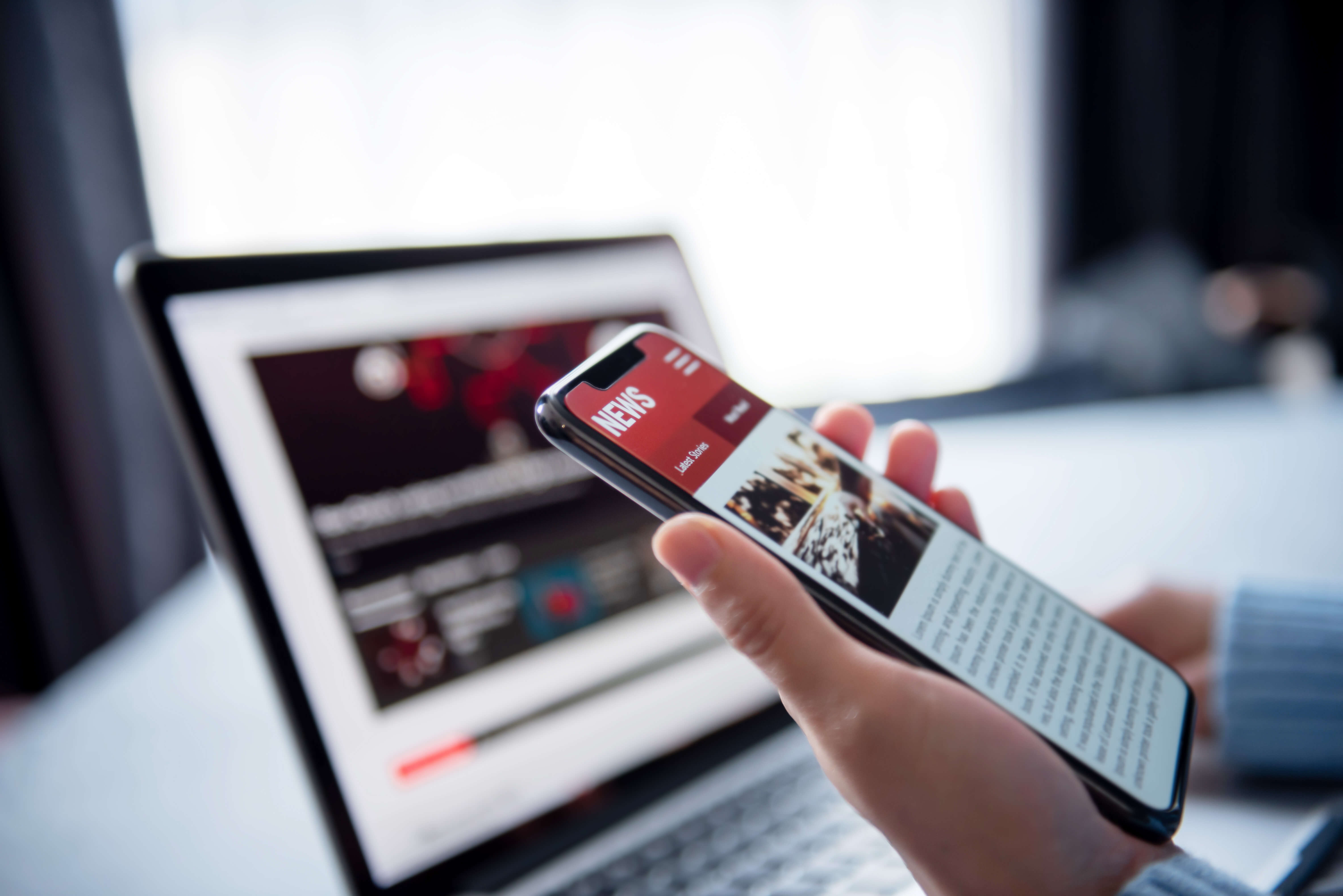 in the foreground a left hand holds a smartphone. its screen reads "News". In the background in soft focus is an open laptop