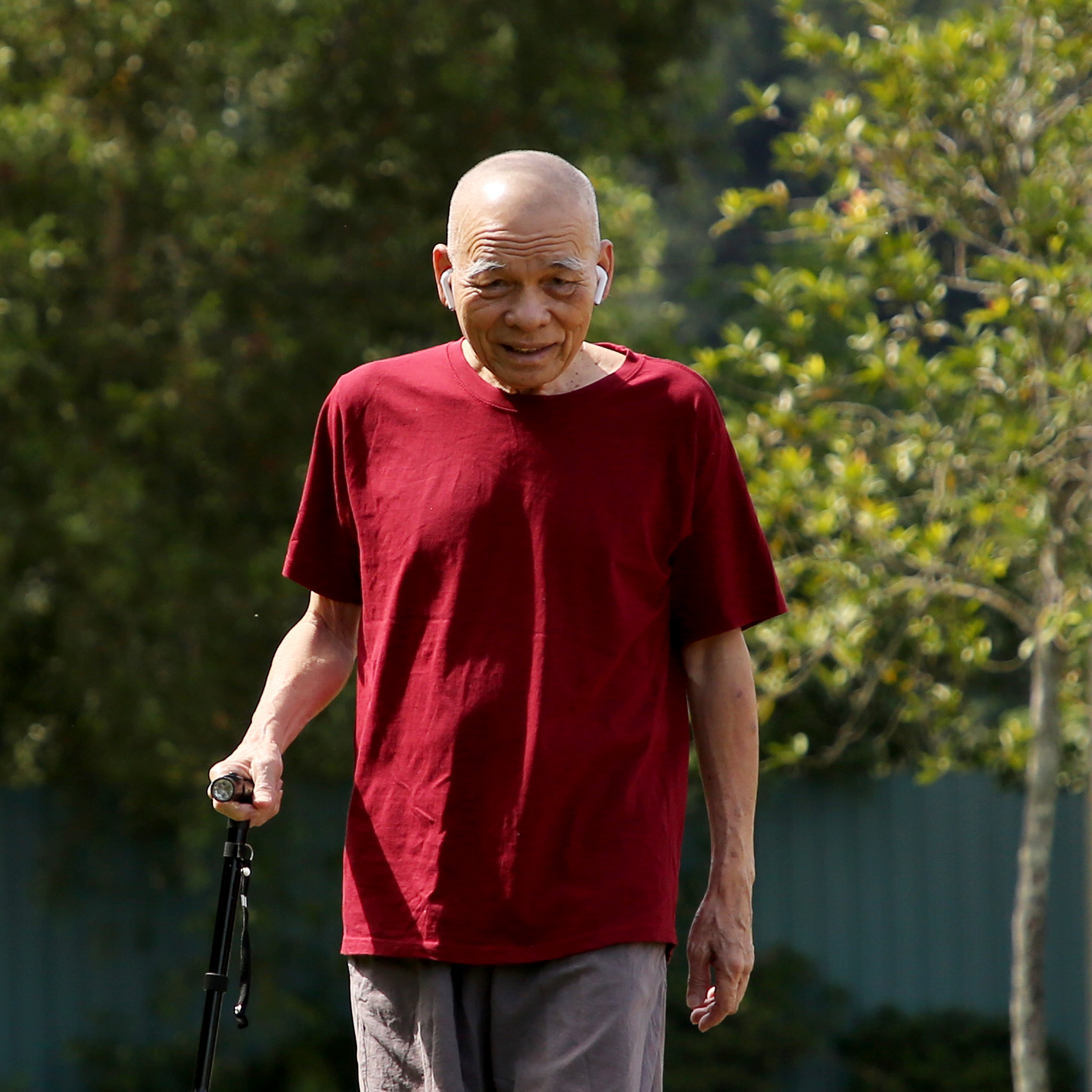 Older man in a deep red tee shirt and earbuds uses a cane to walk a path